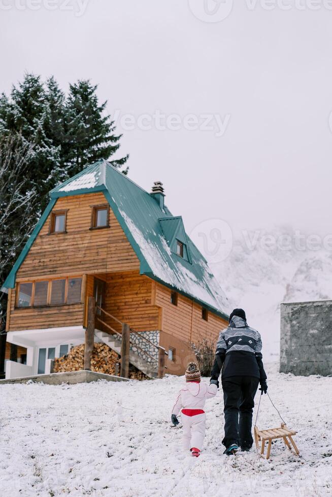 Mother with a sleigh on a rope and a little girl walk up the hill to the cottage, holding hands. Back view photo