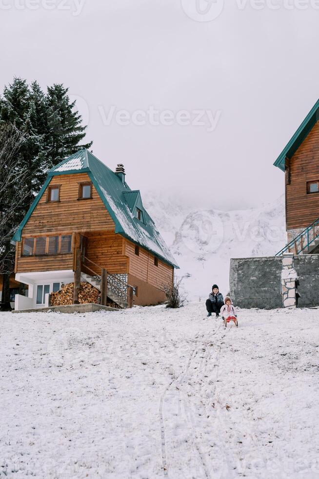 Mom is squatting near the cottage watching a little girl going down the hill on a sled photo