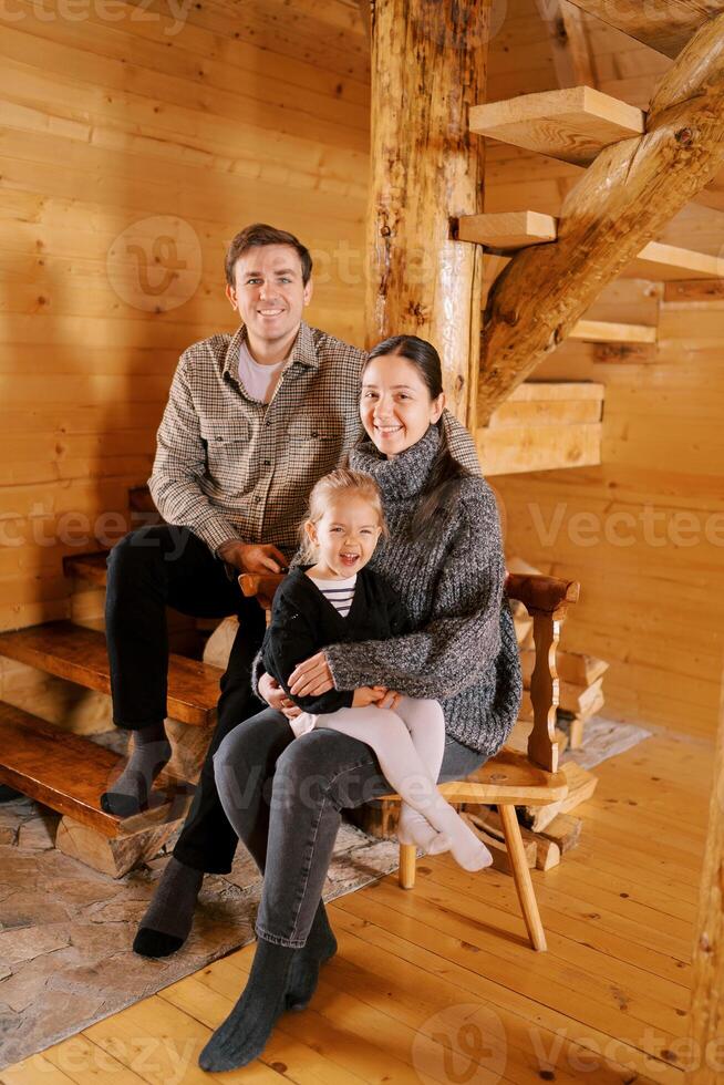 Smiling dad sitting on the stairs next to mom and little girl sitting on a chair in a wooden cottage photo