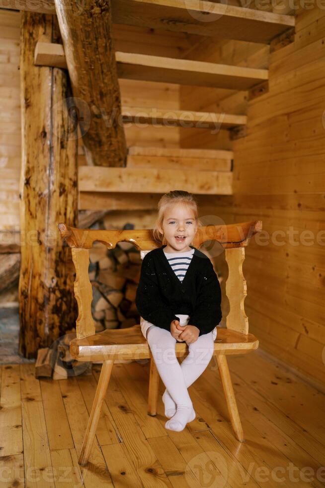 Little laughing girl sitting on a chair near the stairs in a wooden cottage photo