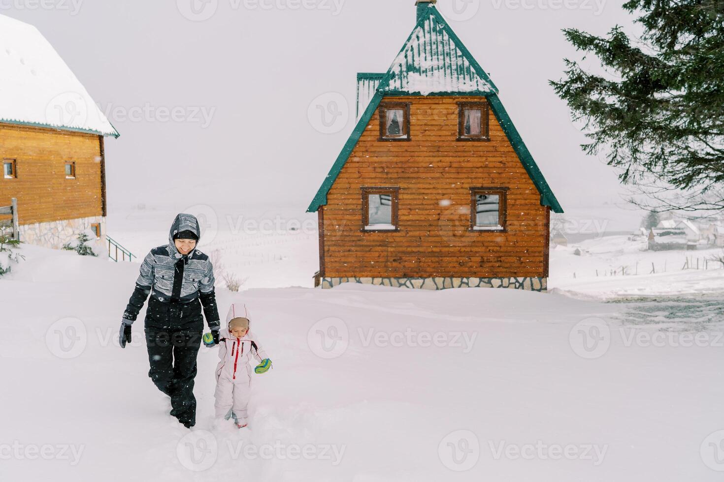 Mom and a little girl walk holding hands through the snowdrifts near wooden houses in the village photo