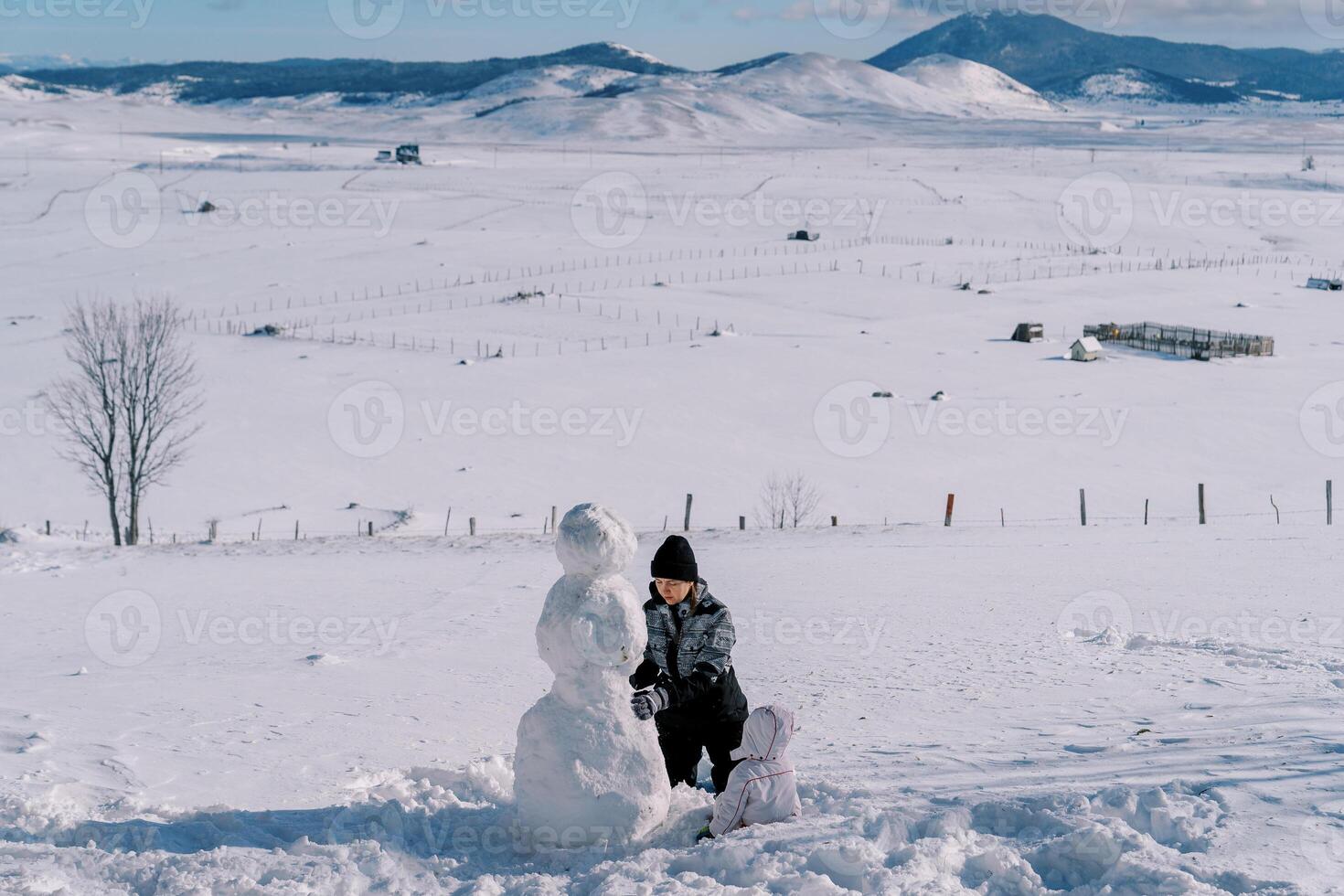 Small child sits in the snow and looks at his mother making a snowman. Back view photo