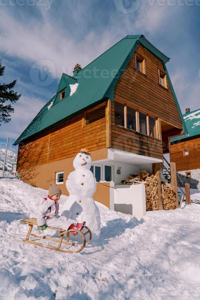Little girl sits on a wooden sled next to a snowman near a wooden cottage. Side view photo