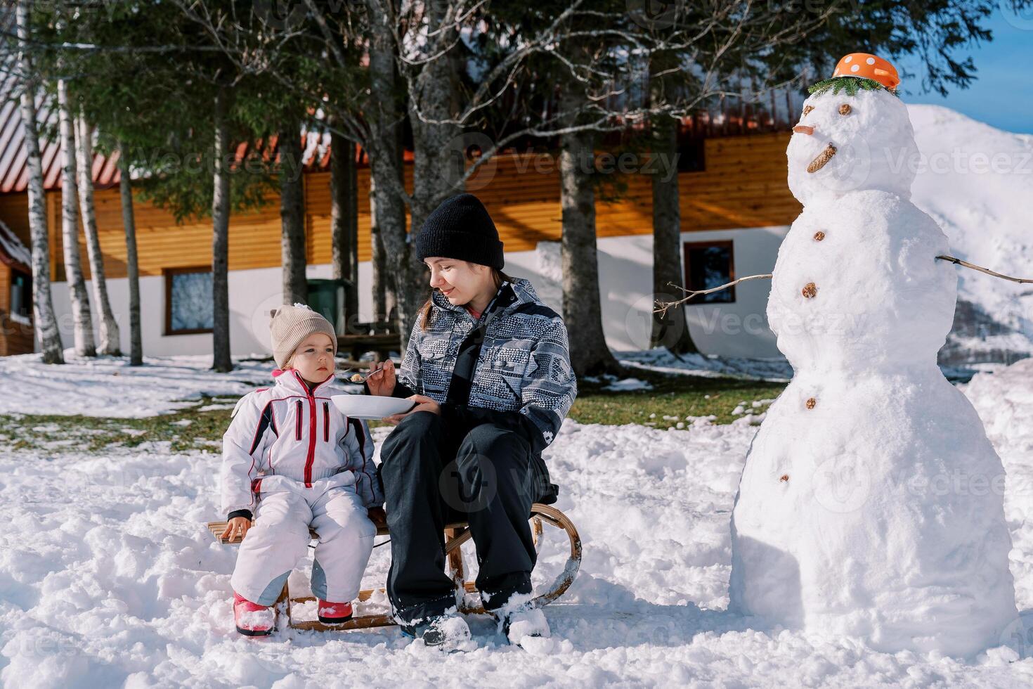 Mom feeds a little girl porridge from a spoon, sitting on a sleigh near a snowman in the yard photo