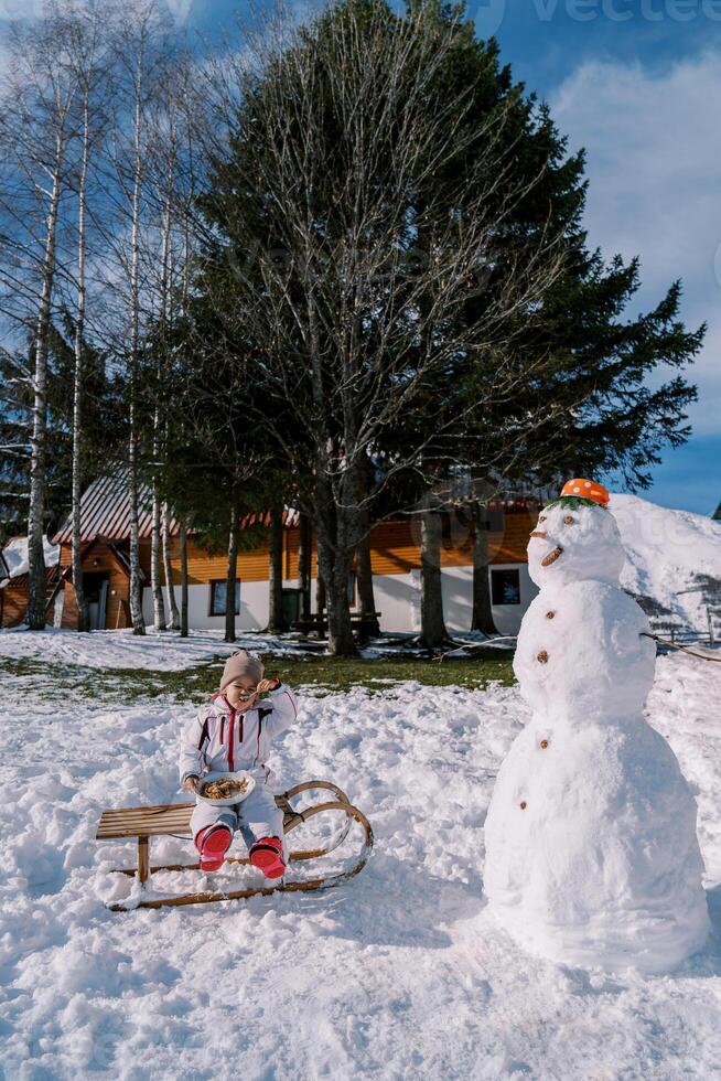 pequeño niña come gachas de avena con un cuchara desde un plato en un trineo cerca un monigote de nieve en el yarda foto