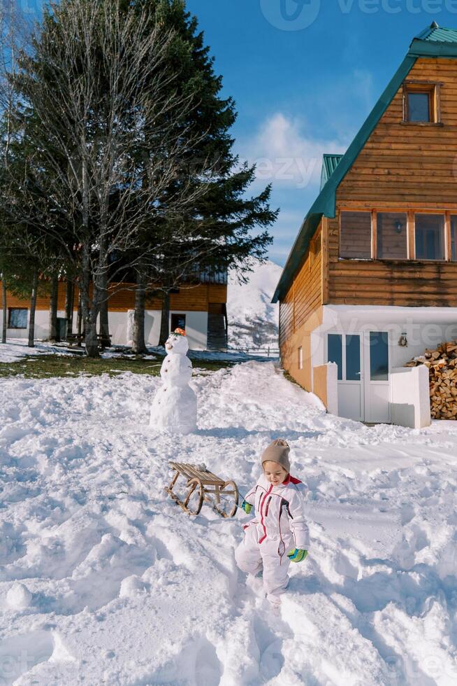 pequeño niña arrastra un trineo en un cuerda desde un colina cerca un de madera chalet foto