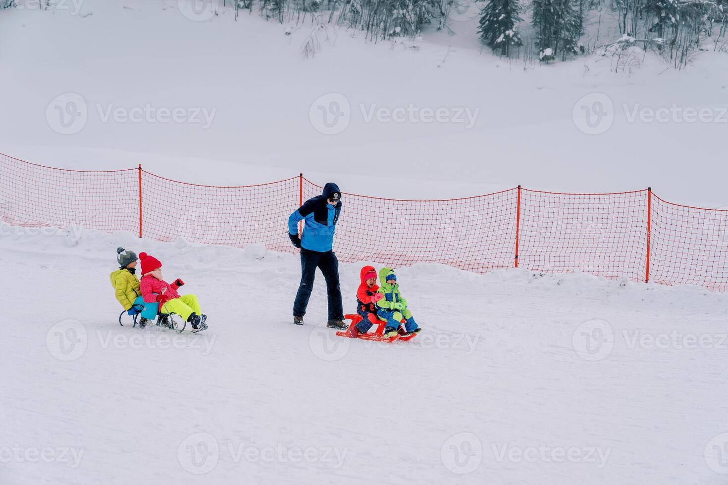 Dad watches children in ski suits riding two by two on sleds down a hill photo