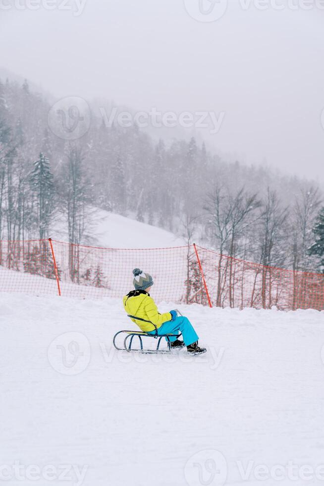 Little boy in a colorful ski suit rides a sled down the slope of a snowy mountain. Side view photo