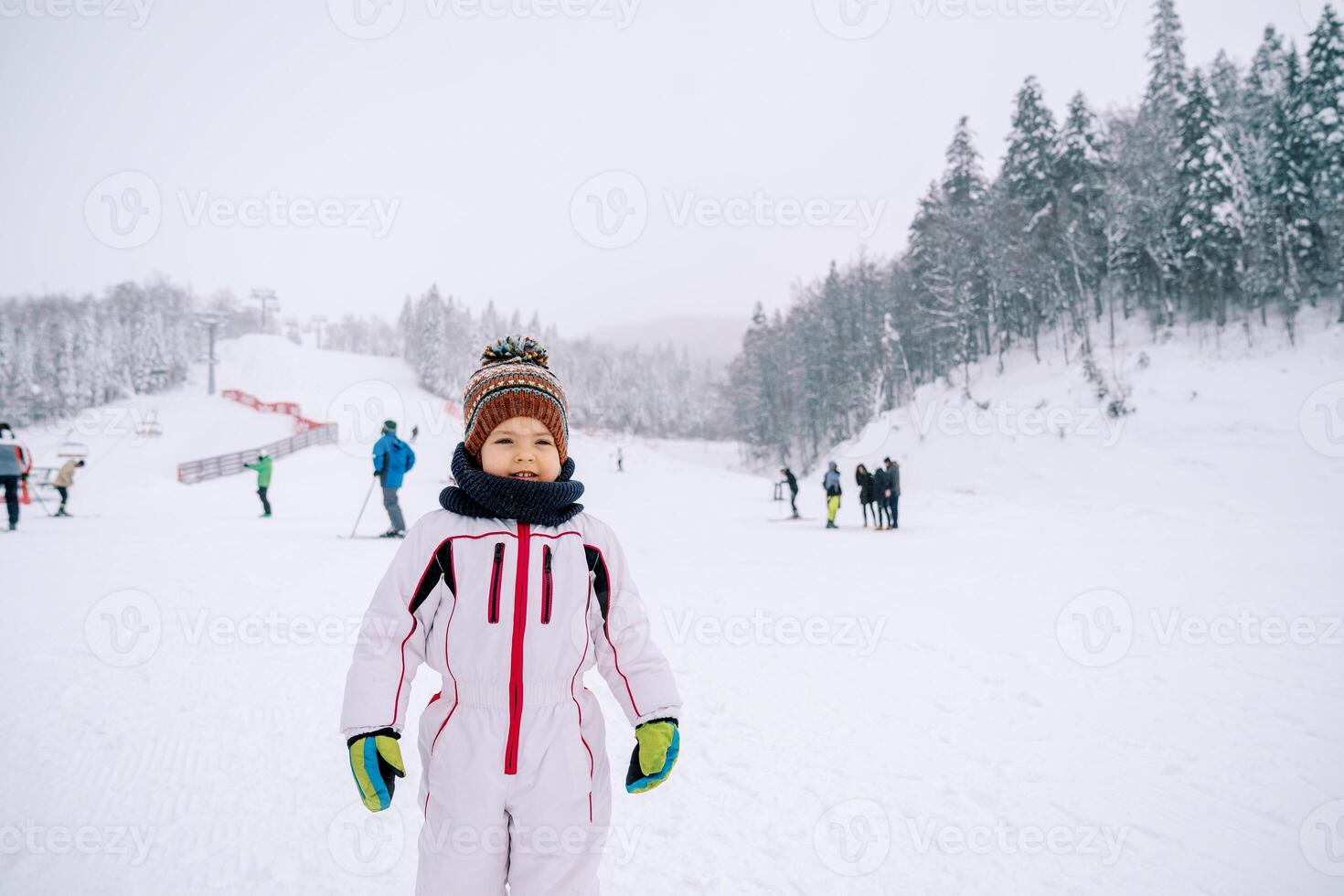 Little girl in a ski suit stands on a ski slope against the backdrop of skiers photo