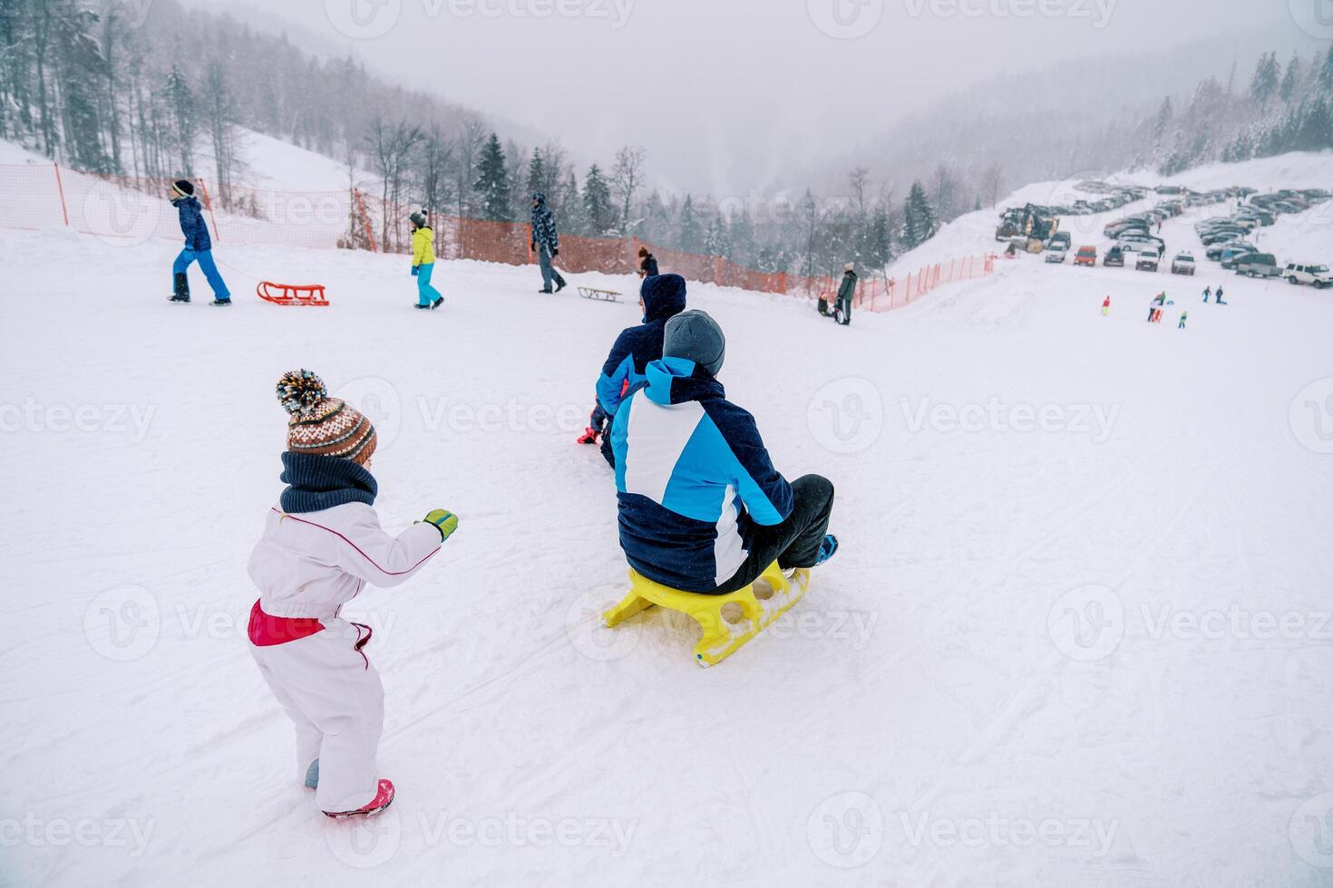 Small child pushes his father on a sled down a snowy hill and looks after him. Back view photo