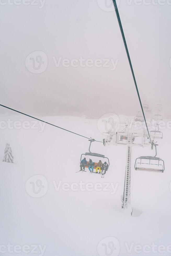 Tourists in colorful ski suits ride uphill through the fog on a chairlift above a snowy forest photo