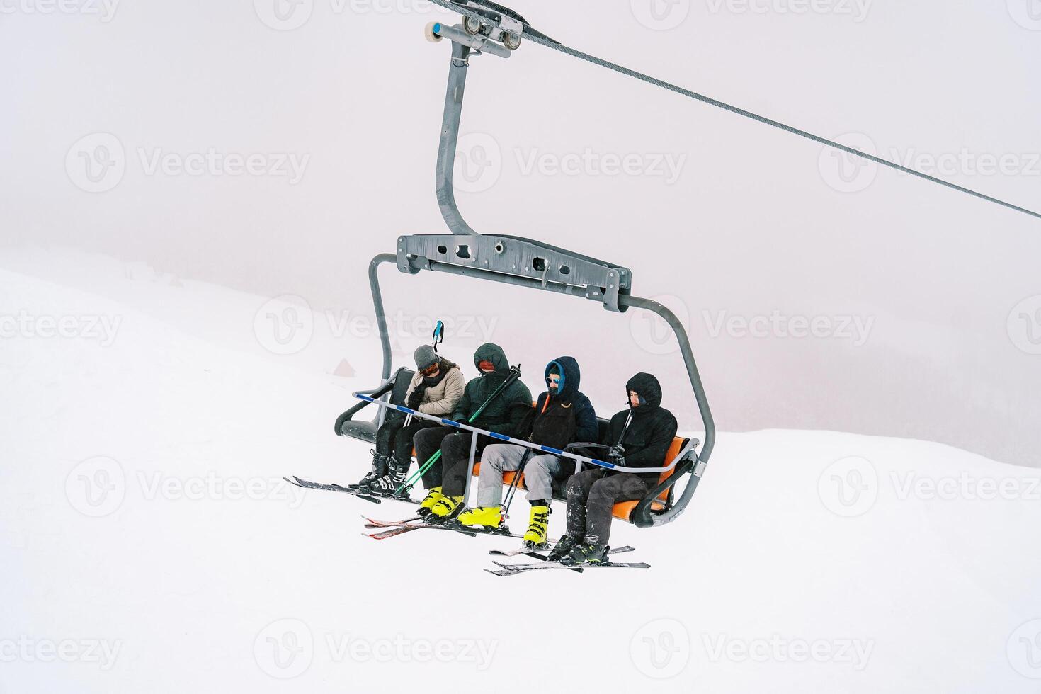 Skiers with ski poles in their hands ride a four-seater chairlift up a snowy mountain slope photo