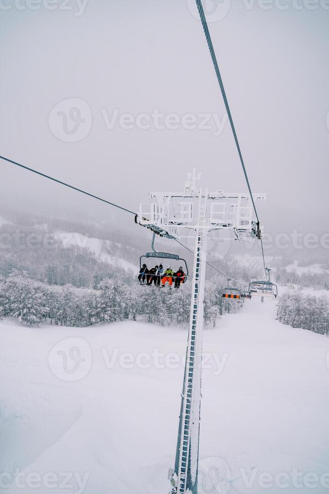 Tourists in colorful ski suits ride on a chairlift over a snowy forest uphill photo