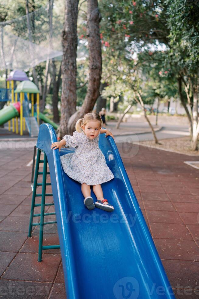 Little girl sits on the top of a slide holding the railing and looking to the side photo