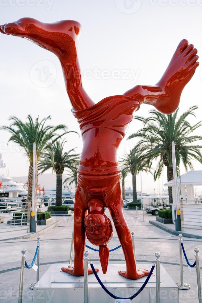 Red sculpture of a girl with large feet standing on her hands on the embankment. Idan Zareski, La Nena photo