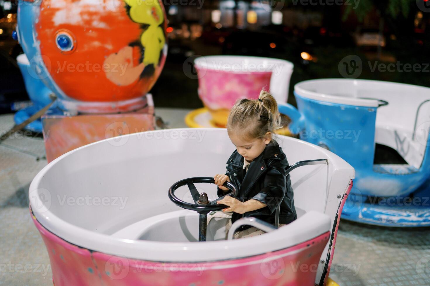 Little girl sits at the wheel of a cup-shaped carousel and looks at her feet photo