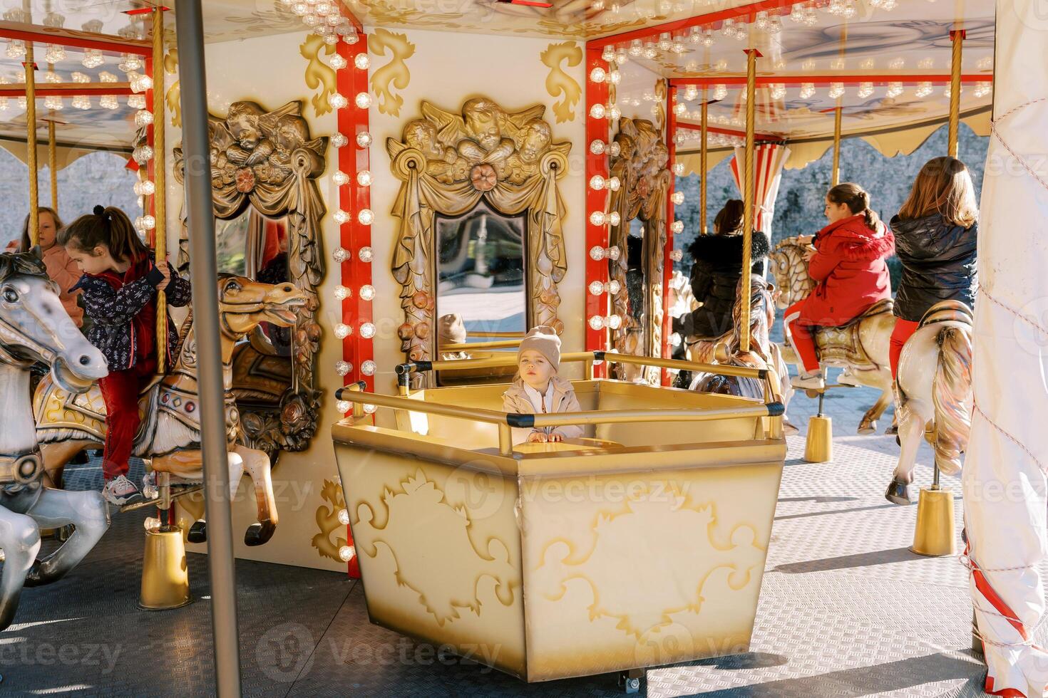 Little girl sits on a carousel seat next to children riding toy horses photo