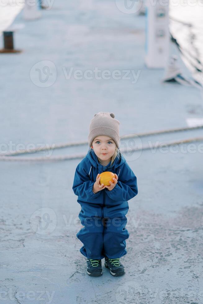 Little girl in overalls with a ripe persimmon in her hands squatted on the road photo