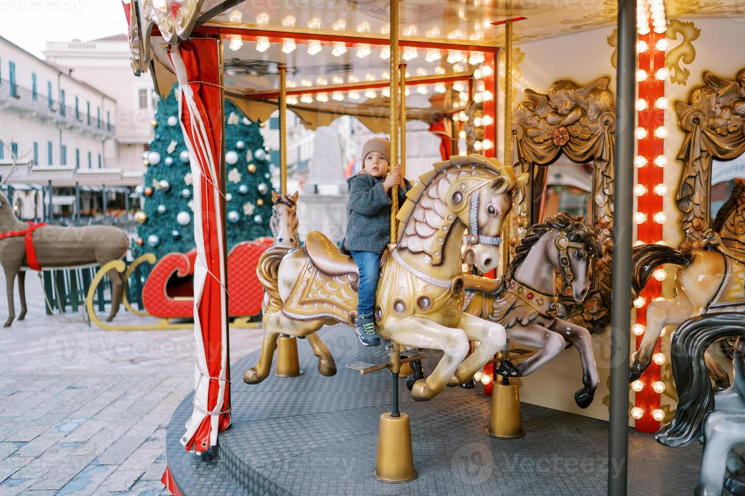 Little girl rides a toy horse on a carousel at a fair near a decorated Christmas tree near an old house photo