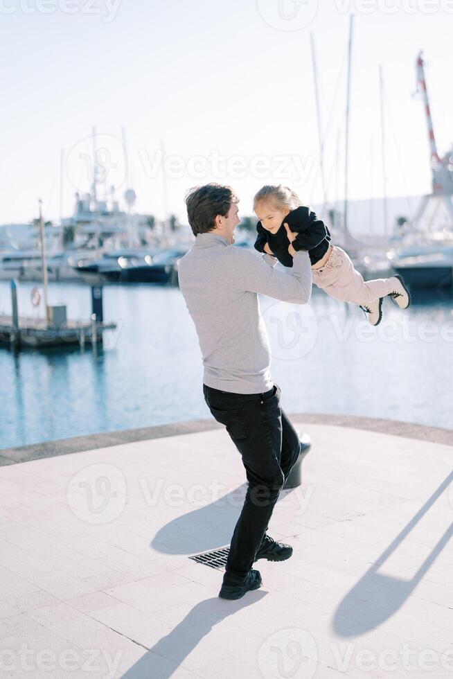 Smiling dad spins a little girl in his arms on the pier. Back view photo