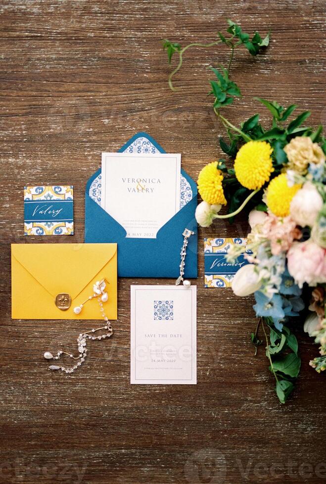 Wedding invitations with blue and yellow envelopes, name cards and a bouquet of flowers lie on a wooden table photo