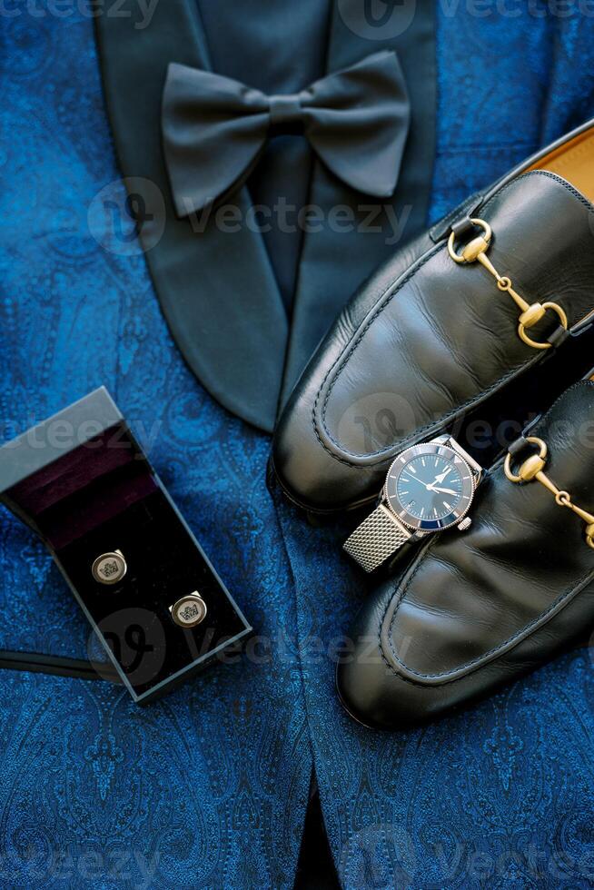 Black men shoes stand on a blue suit next to a watch and cufflinks in a box photo