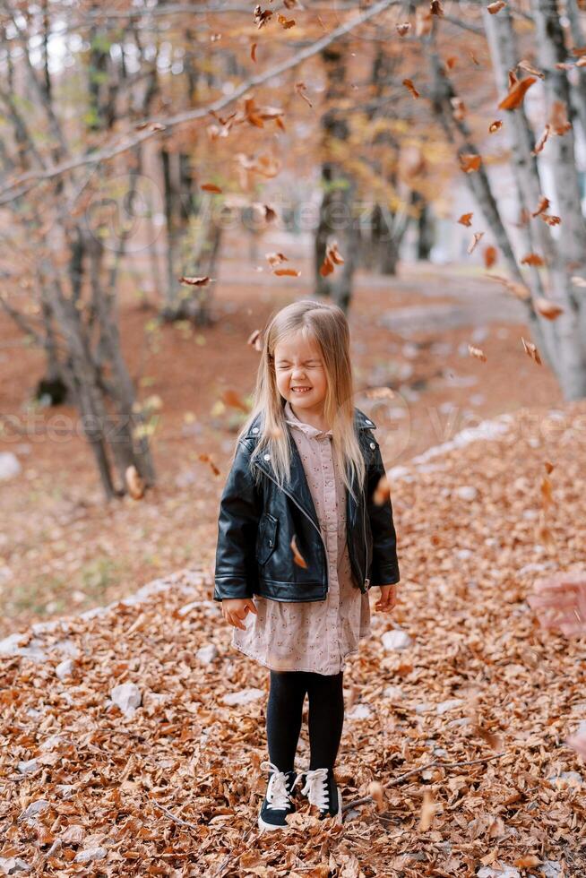 Little girl stands with her eyes closed under falling dry leaves in the autumn forest photo