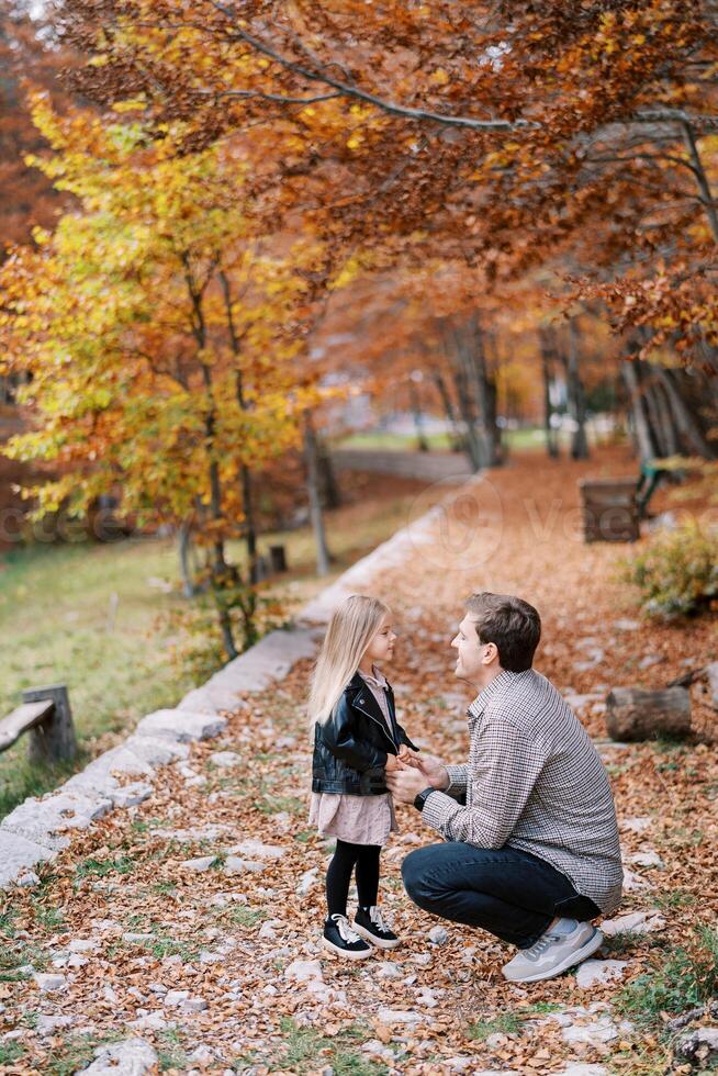 papá en cuclillas en frente de el pequeño niña participación su manos en un rocoso camino en el otoño bosque foto
