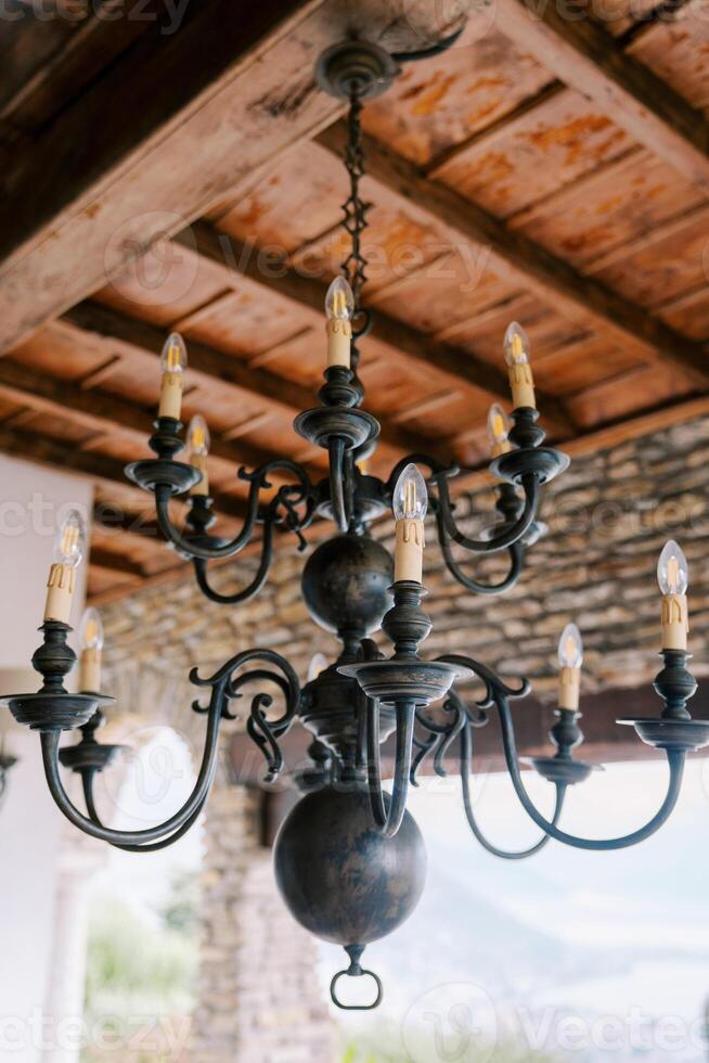 Bronze branched chandelier with spheres and candle bulbs hangs from the wooden ceiling of an ancient villa photo