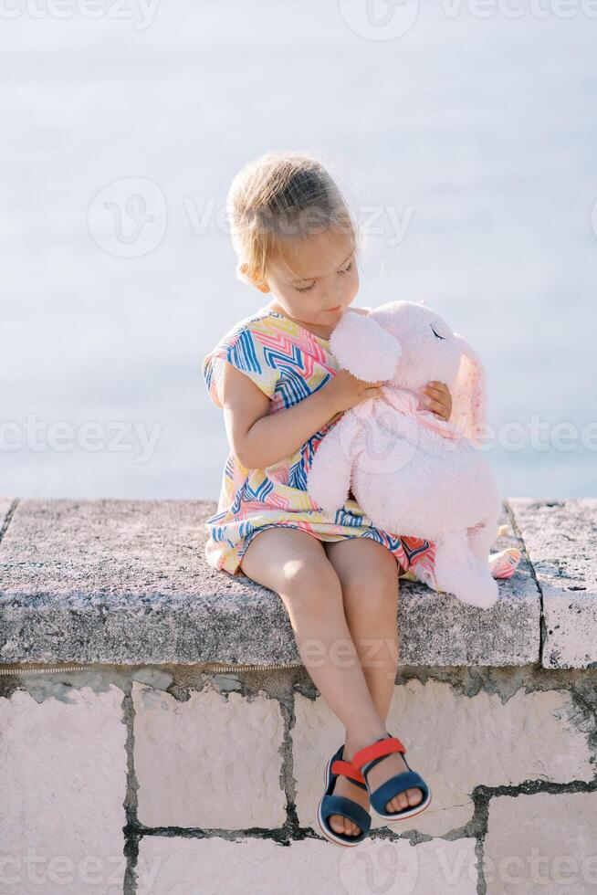 Little girl examines a plush rabbit in her hands while sitting on a fence by the sea photo