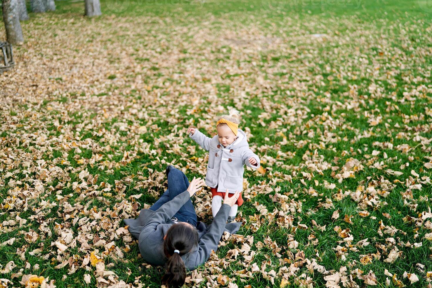 Mom lying on dry foliage stretches out her hands to a little girl standing on a green lawn photo