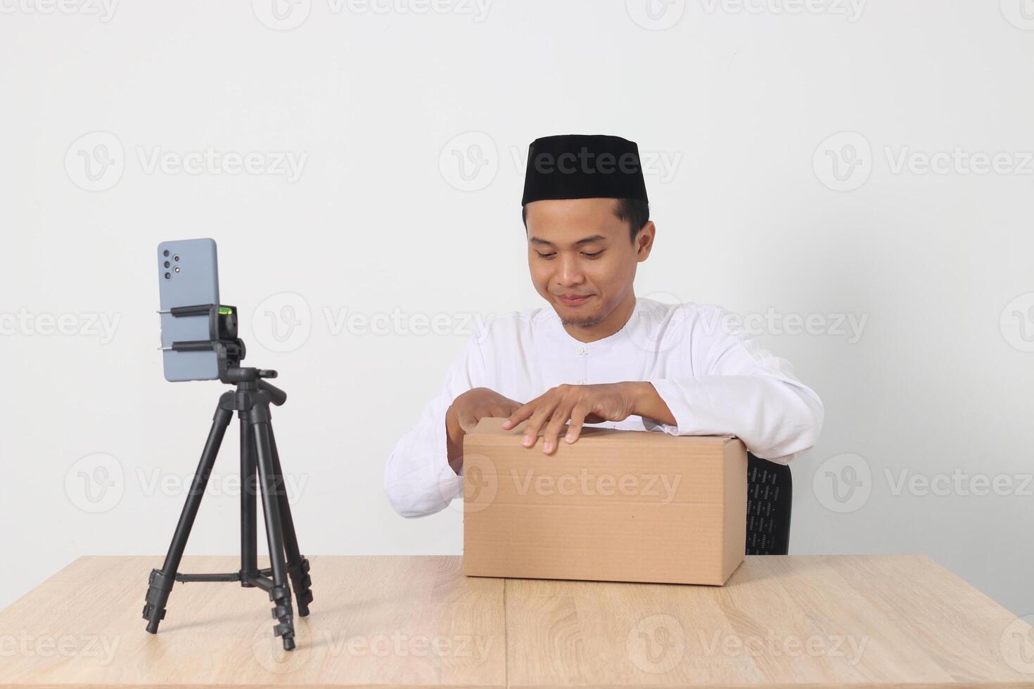 Portrait of excited Asian muslim man in koko shirt with skullcap promoting his product on live streaming session. Online shopping with smartphone concept. Isolated image on white background photo