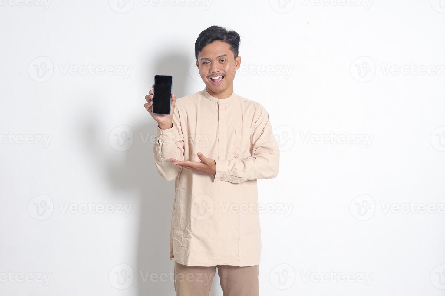 Portrait of young excited Asian muslim man in koko shirt showing blank screen mobile phone mockup while pointing and presenting product. Social media concept. Isolated image on white background photo