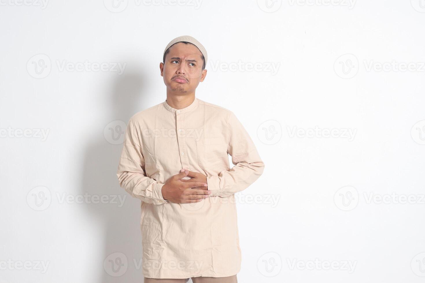 Portrait of religious Asian muslim man in koko shirt with skullcap feeling pain in his stomach, endure hunger. Stomachache and hungry for food concept. Isolated image on white background photo