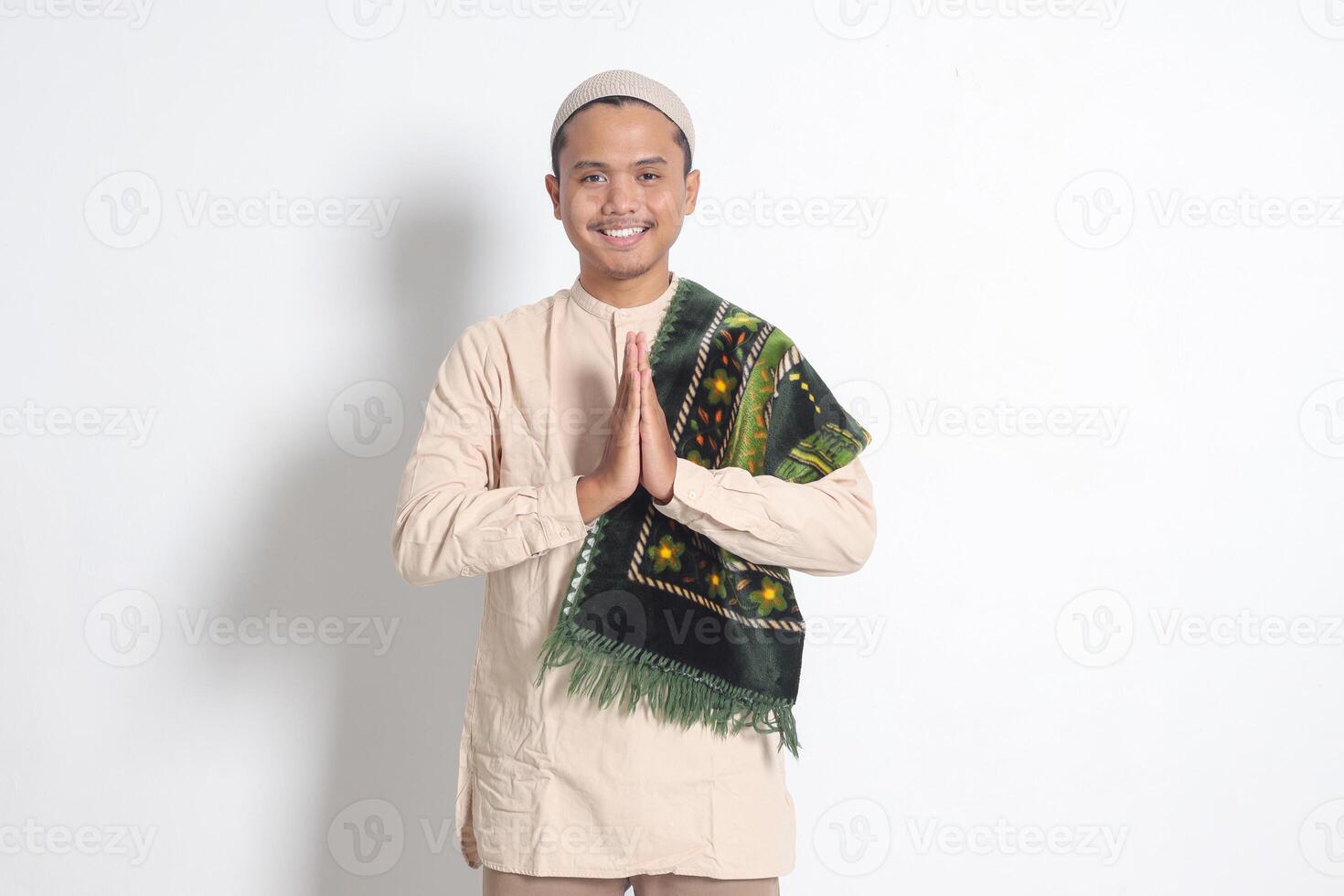 Portrait of attractive Asian muslim man in koko shirt with prayer mat showing apologize and welcome hand gesture. Apology during eid mubarak. Isolated image on white background photo