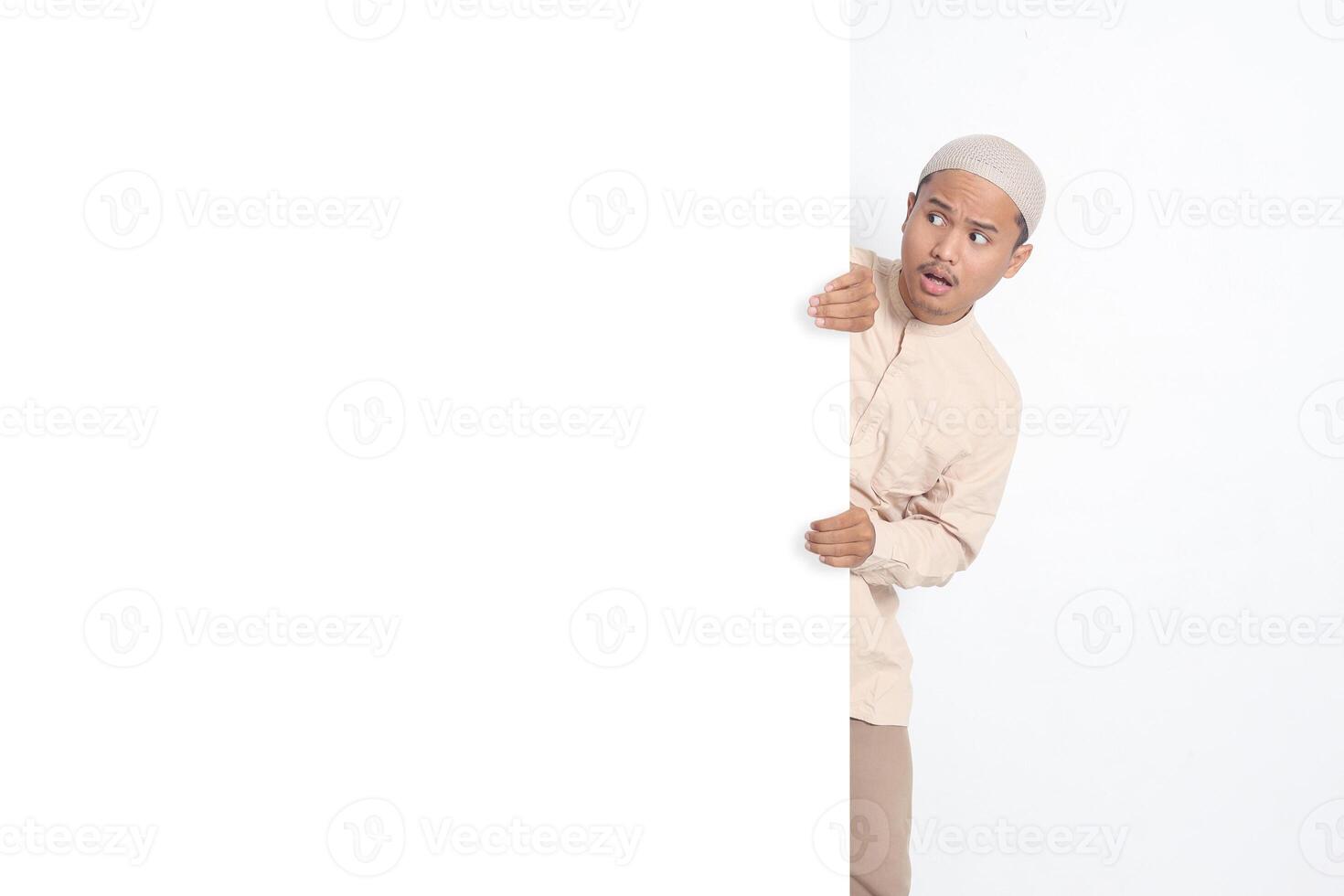 Portrait of excited Asian muslim man in koko shirt hiding behind empty white billboard with mockup template and copy space for graphic design. Advertising poster. Isolated image on white background photo