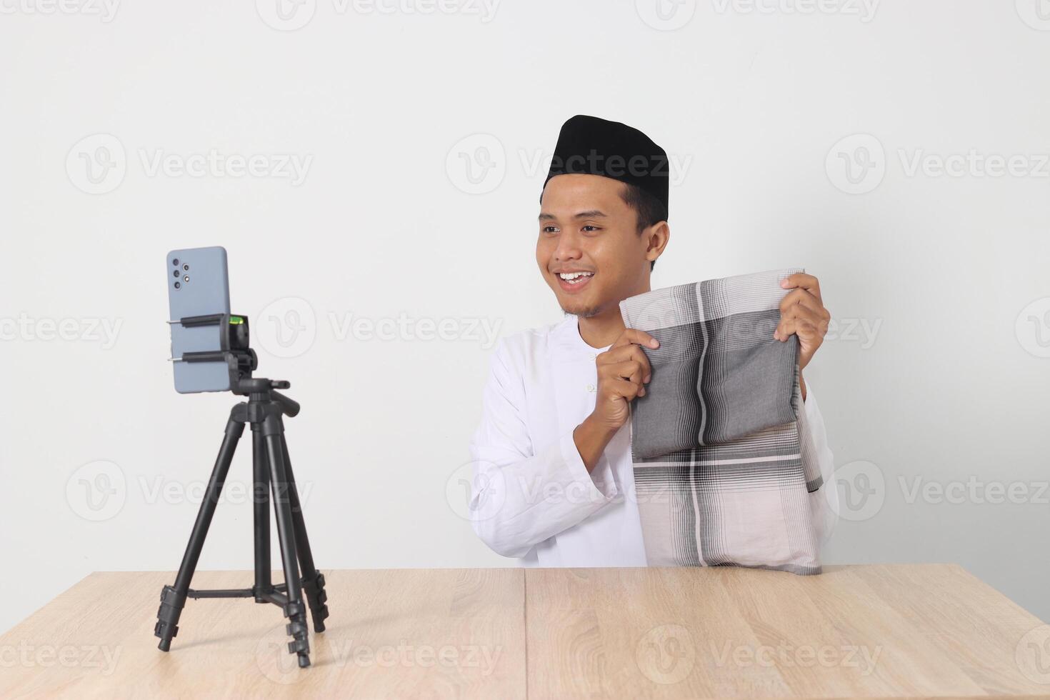 Portrait of excited Asian muslim man in koko shirt with skullcap promoting his product, sarung, on live streaming session. Online shopping with smartphone concept. Isolated image on white background photo