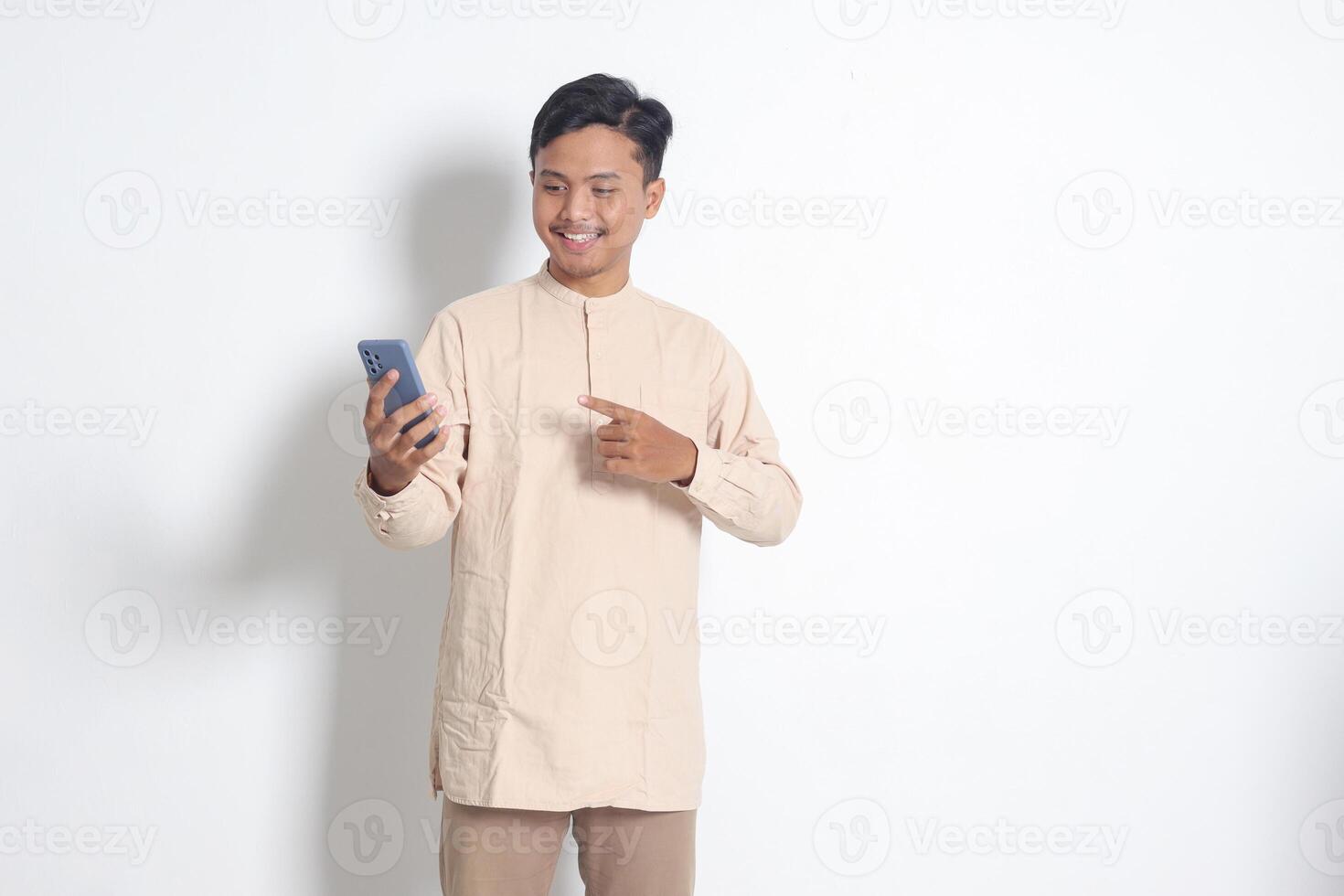 Portrait of young excited Asian muslim man in koko shirt holding mobile phone, pointing and showing product with finger. Social media concept. Isolated image on white background photo