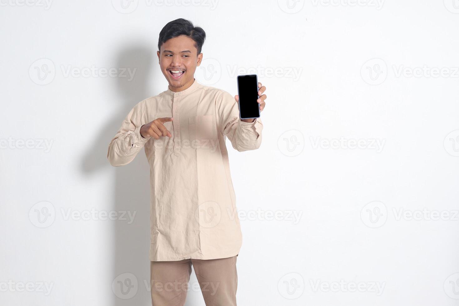 Portrait of young excited Asian muslim man in koko shirt showing blank screen mobile phone mockup while pointing and presenting product. Social media concept. Isolated image on white background photo