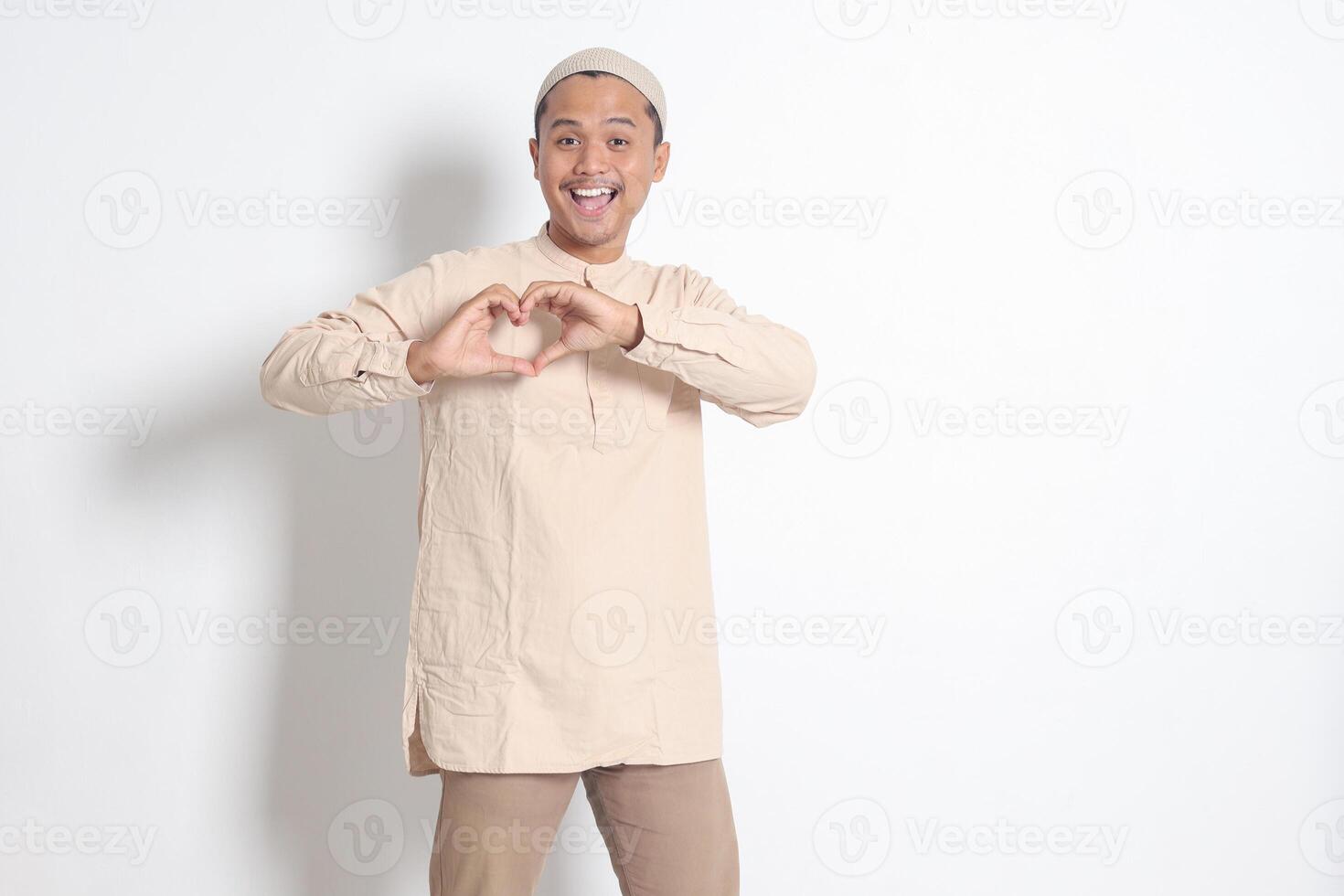 Portrait of excited Asian muslim man in koko shirt with skullcap speaks own feelings, makes heart gesture over chest, expresses sympathy and love, smiles positively. Isolated image on white background photo
