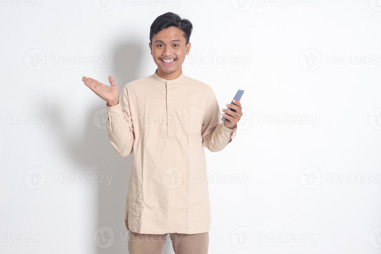 Portrait of young excited Asian muslim man in koko shirt holding mobile phone, pointing and showing product with finger. Social media concept. Isolated image on white background photo