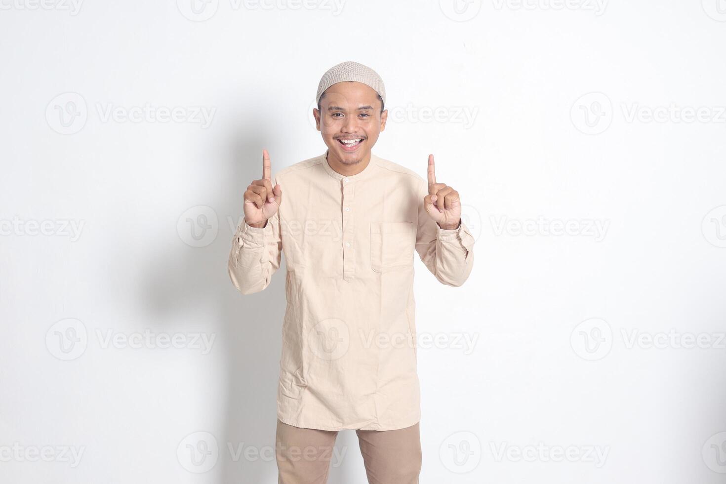 Portrait of shocked Asian muslim man in koko shirt with skullcap showing product and pointing with his hand and finger up. Advertising concept. Isolated image on white background photo