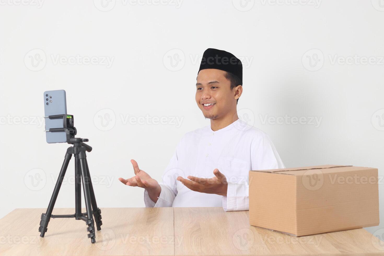 Portrait of excited Asian muslim man in koko shirt with skullcap promoting his product on live streaming session. Online shopping with smartphone concept. Isolated image on white background photo