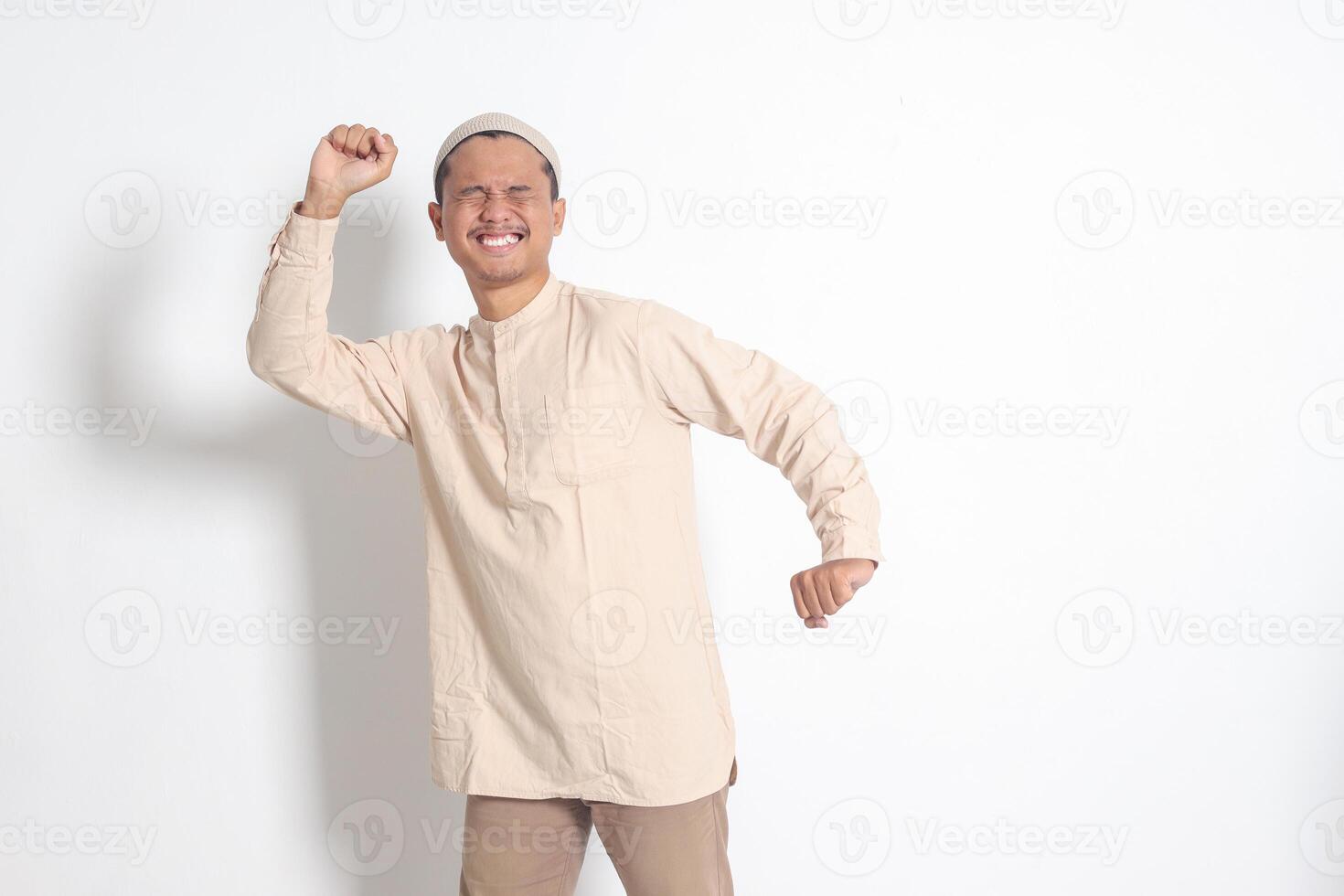 Portrait of overworked Asian muslim man in koko shirt with skullcap stretching his hands and body after waking up. Sleep deprivation concept. Isolated image on white background photo