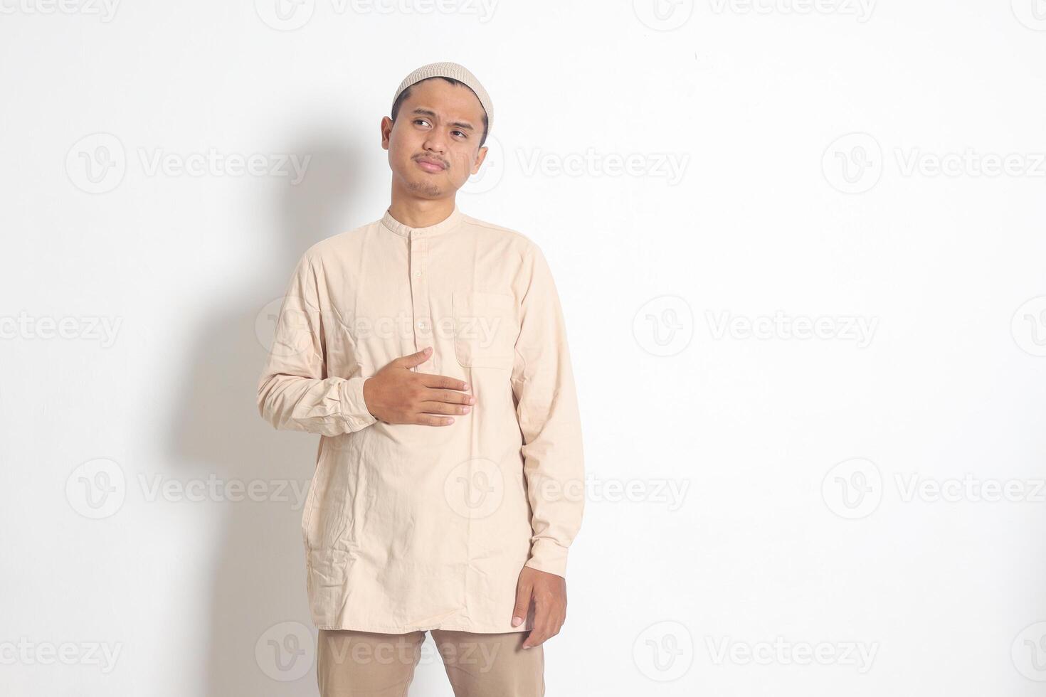 Portrait of religious Asian muslim man in koko shirt with skullcap feeling pain in his stomach, endure hunger. Stomachache and hungry for food concept. Isolated image on white background photo