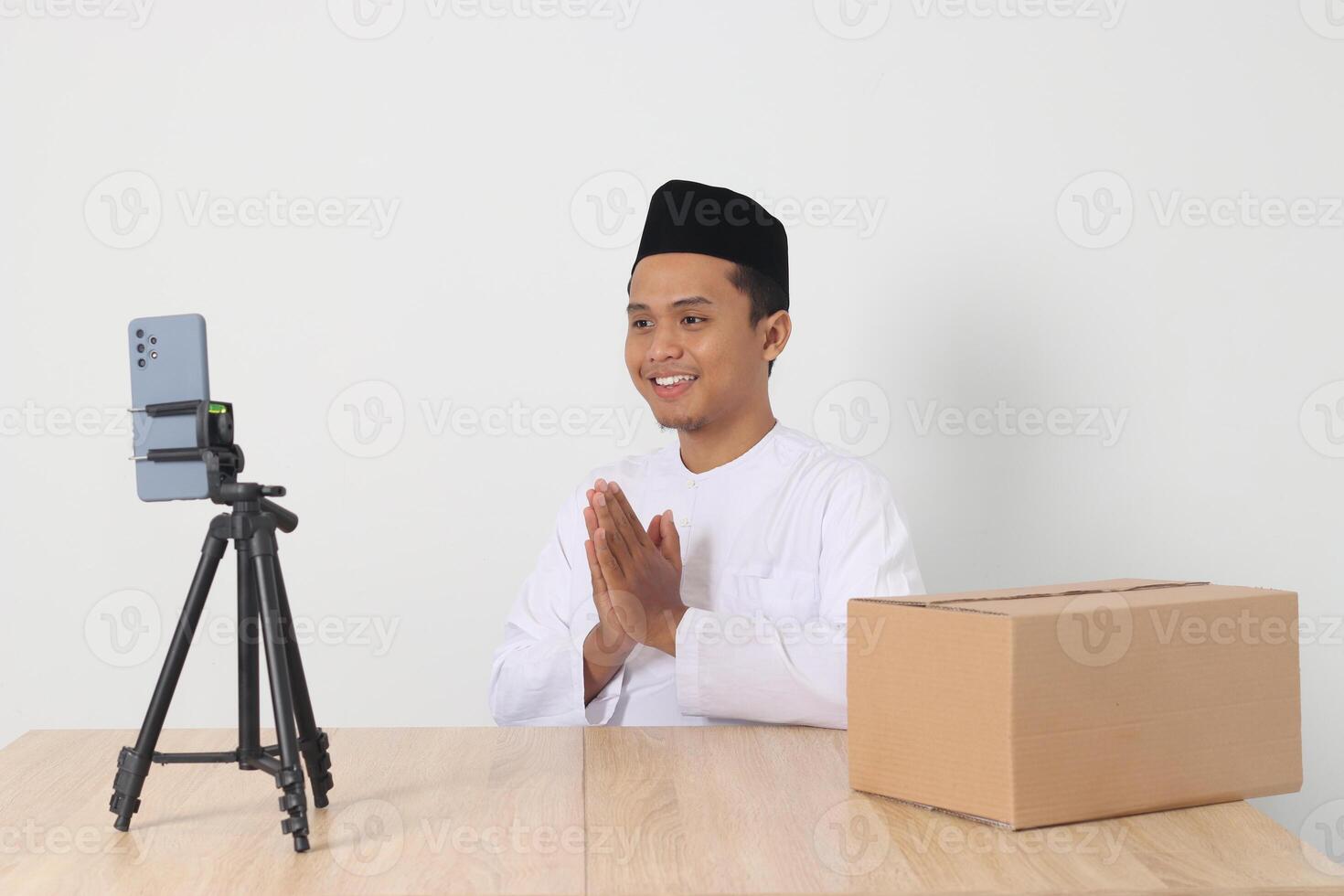 Portrait of excited Asian muslim man in koko shirt with skullcap promoting his product on live streaming session. Online shopping with smartphone concept. Isolated image on white background photo