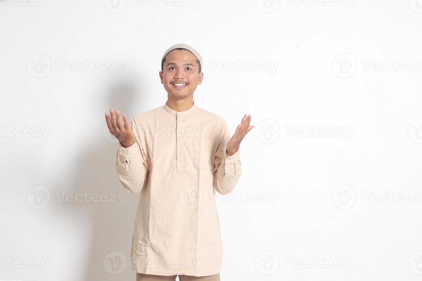 Portrait of religious Asian muslim man in koko shirt with skullcap praying earnestly with his hands raised. Devout faith concept. Isolated image on white background photo
