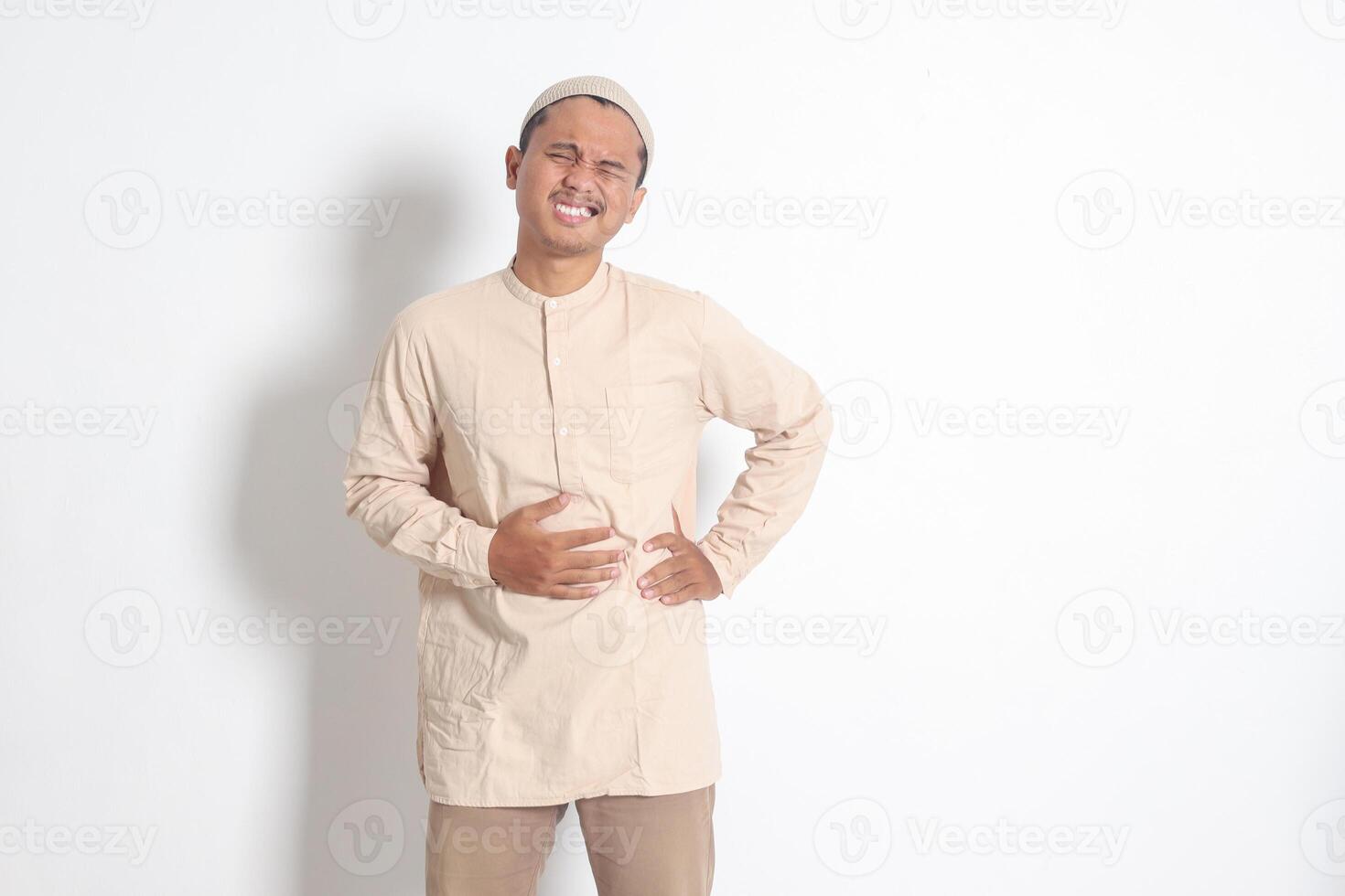 Portrait of religious Asian muslim man in koko shirt with skullcap feeling pain in his stomach, endure hunger. Stomachache and hungry for food concept. Isolated image on white background photo