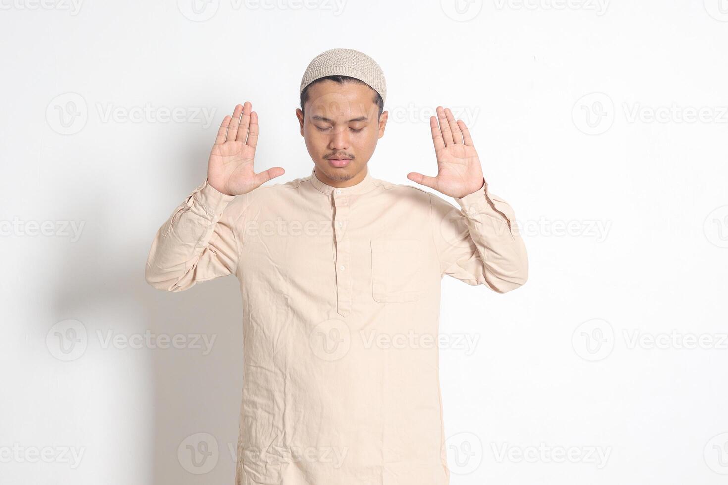 Portrait of religious Asian muslim man in koko shirt with skullcap praying with hands raised, doing takbir sholat before the pray. Isolated image on white background photo