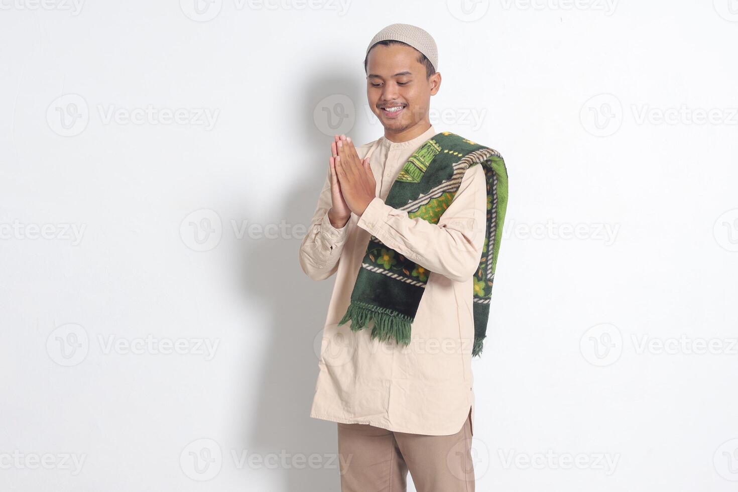 Portrait of attractive Asian muslim man in koko shirt with prayer mat showing apologize and welcome hand gesture. Apology during eid mubarak. Isolated image on white background photo